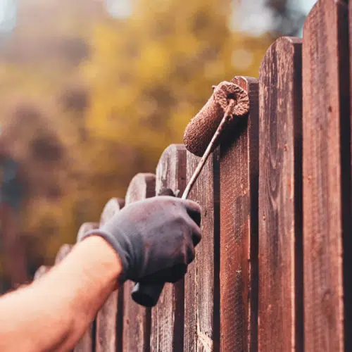 Person painting a fence brown with a paint roller